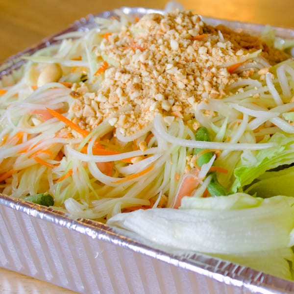 Close-up of a vibrant papaya salad with shredded carrots, green beans, and crushed peanuts in a foil container.
