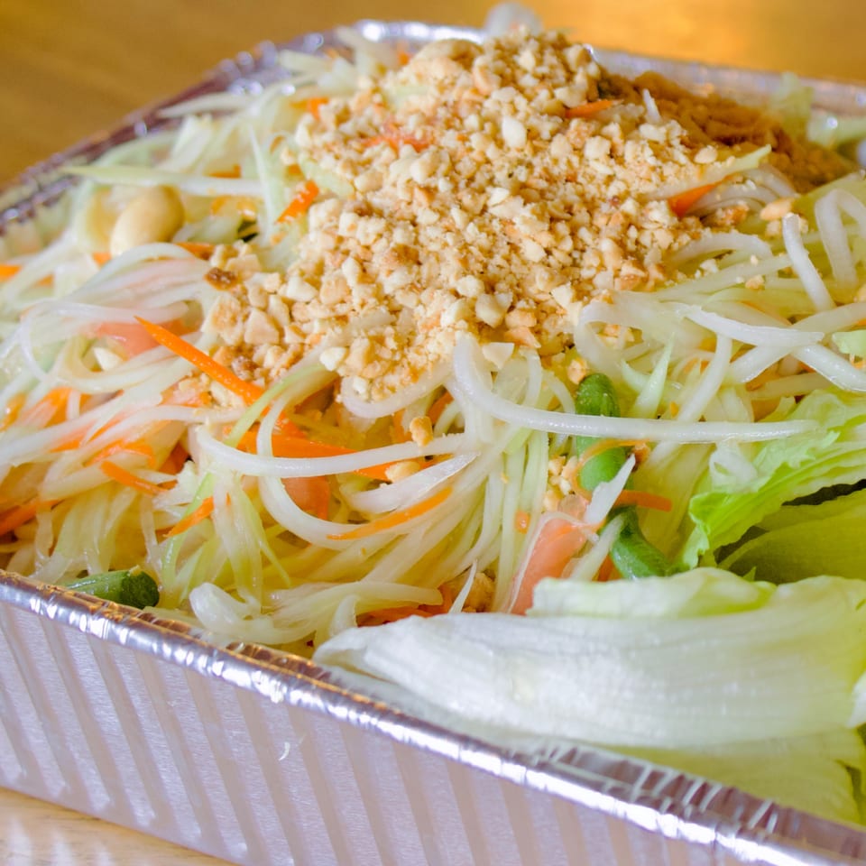 Close-up of a vibrant papaya salad with shredded carrots, green beans, and crushed peanuts in a foil container.