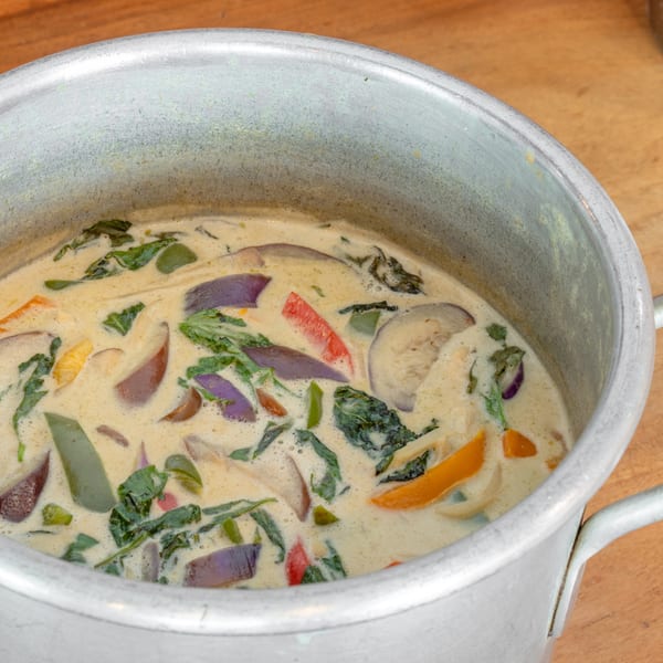 Creamy green curry with eggplant, bell peppers, and leafy greens in a metal pot.