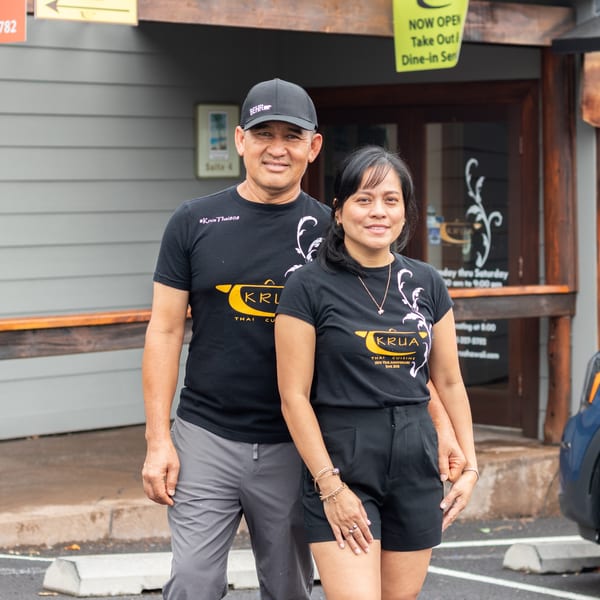 Owners of Krua Thai Cuisine, a man and woman, stand smiling in front of their restaurant.