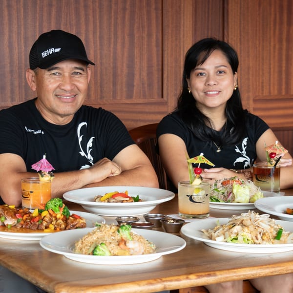 Two smiling people, a man and a woman, seated at a table filled with various Thai dishes and drinks.