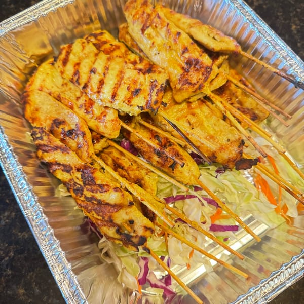 Grilled chicken satay skewers with char marks, served over shredded cabbage in a foil container.
